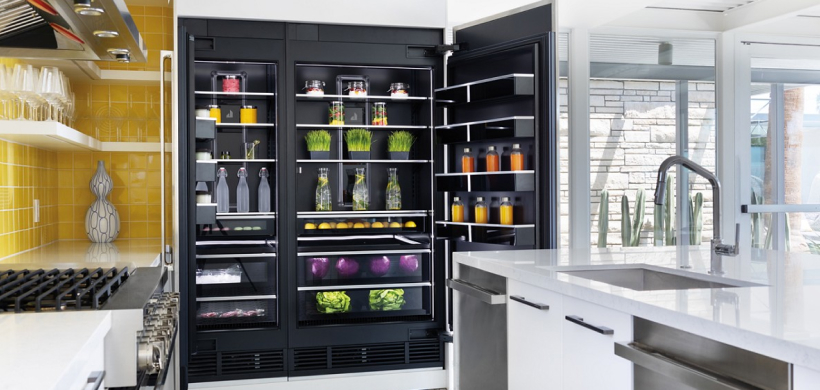 Une cuisine avec une table de cuisson JennAir contre un dosseret jaune. Sur les étagères, on peut voir des verres à vin et un vase sur le comptoir. Un réfrigérateur JennAir ouvert est rempli de différentes boissons, de fruits et légumes, de desserts et d'herbes fraîches.